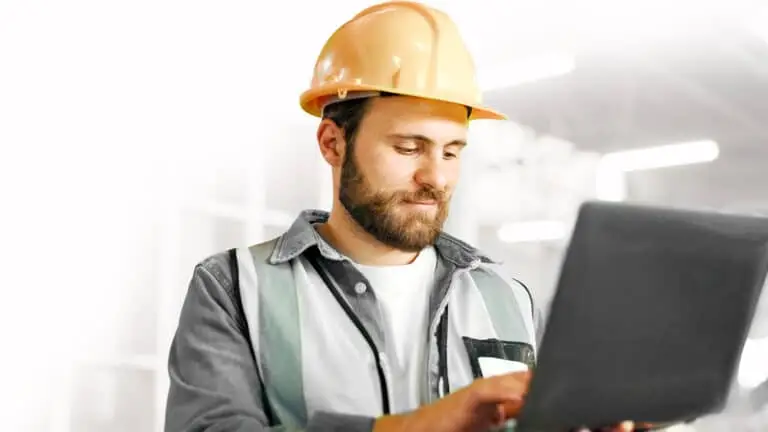 Man wearing a yellow hard hat and safety vest uses a laptop in an indoor construction or industrial setting.