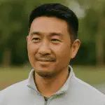 A man with short black hair and a trimmed beard is wearing a light gray zip-up jacket, standing outdoors with greenery blurred in the background.