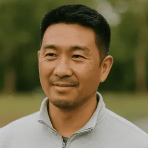 A man with short black hair and a trimmed beard is wearing a light gray zip-up jacket, standing outdoors with greenery blurred in the background.