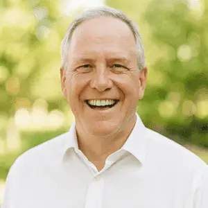 An older man with gray hair, wearing a white shirt, smiles broadly outdoors with greenery in the blurred background.