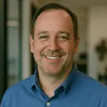 A middle-aged man with short brown hair smiles at the camera. He is wearing a blue collared shirt and is standing indoors in an office setting.