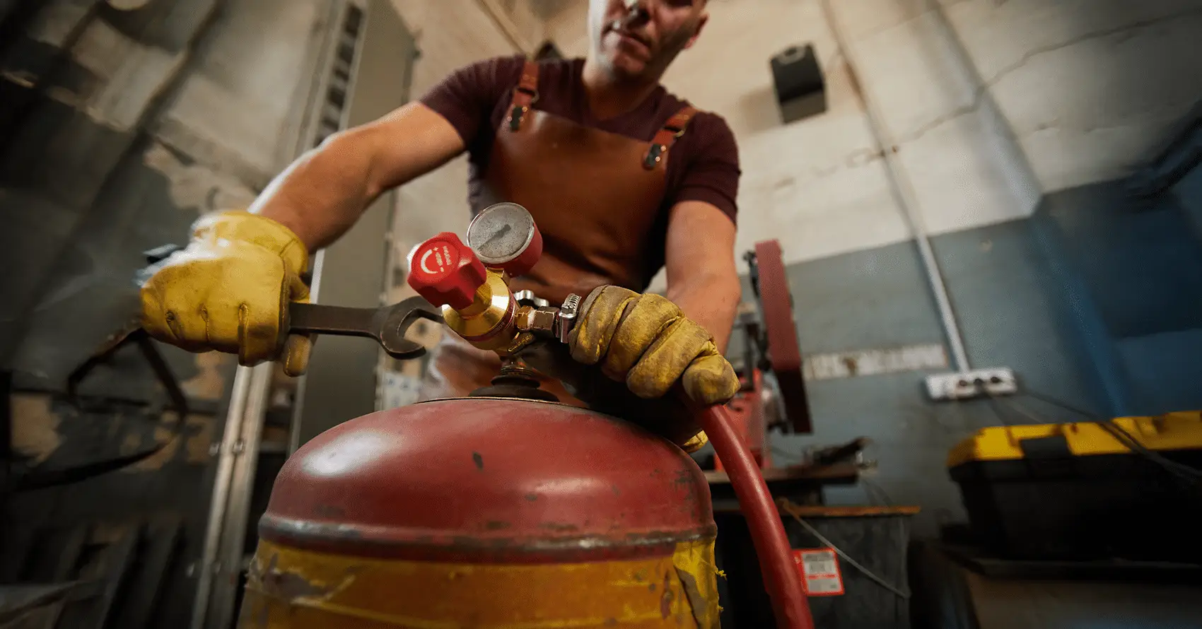 Person wearing gloves and an apron adjusting a valve on a red gas cylinder in an industrial workshop.