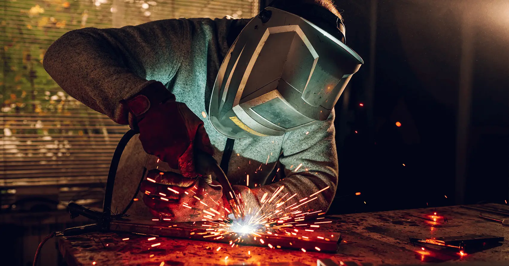 A person wearing a welding helmet and gloves welds metal, with bright sparks flying from the work area on a workshop table.