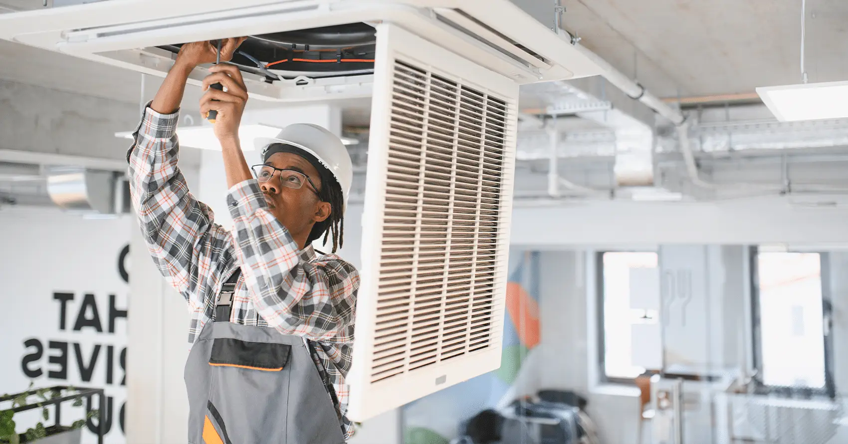 A worker in a hard hat and overalls repairs or installs an overhead air conditioning unit in a modern office space.