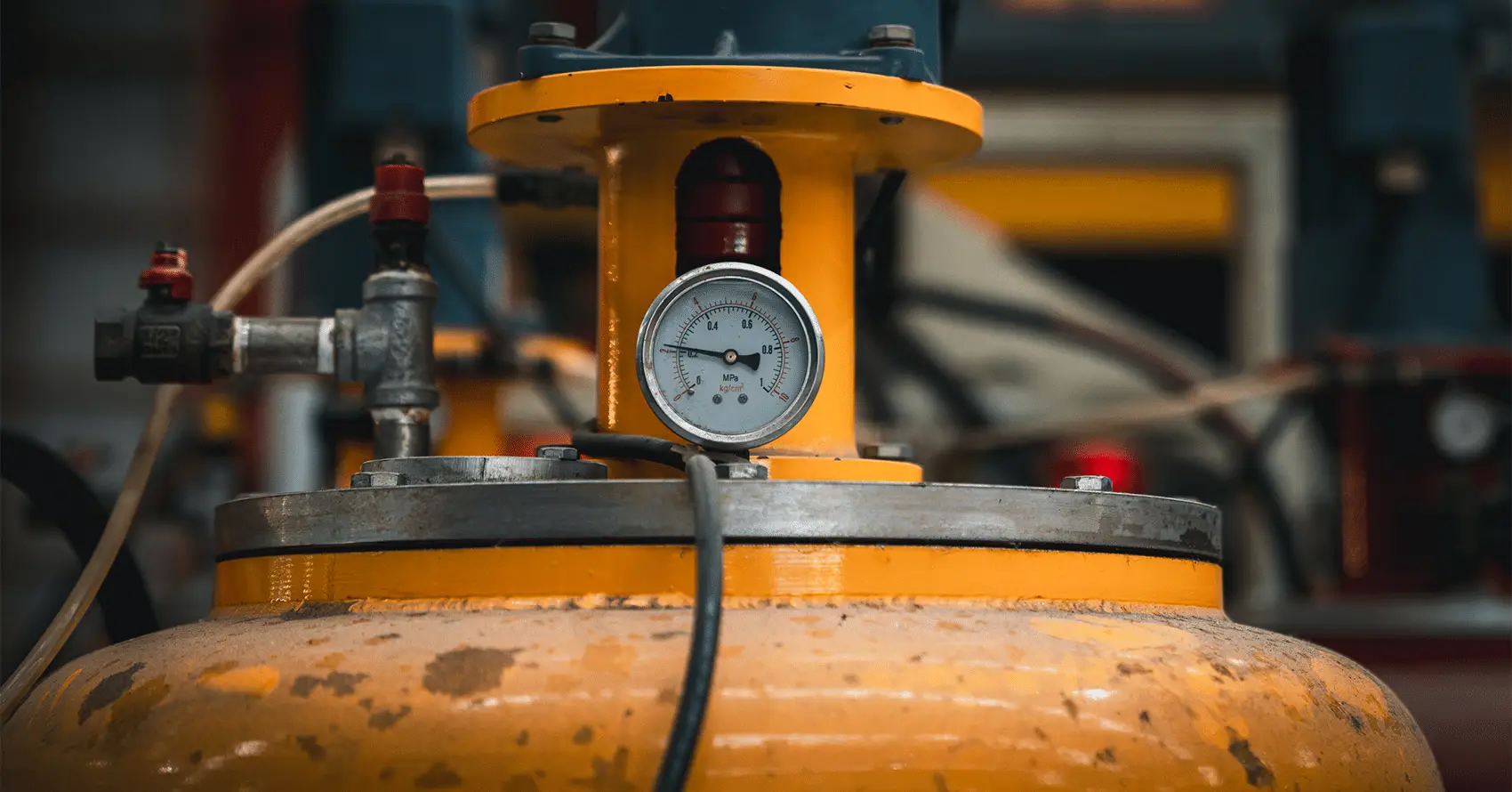 A close-up of a pressure gauge attached to a yellow industrial gas cylinder with pipes and valves visible in the background.