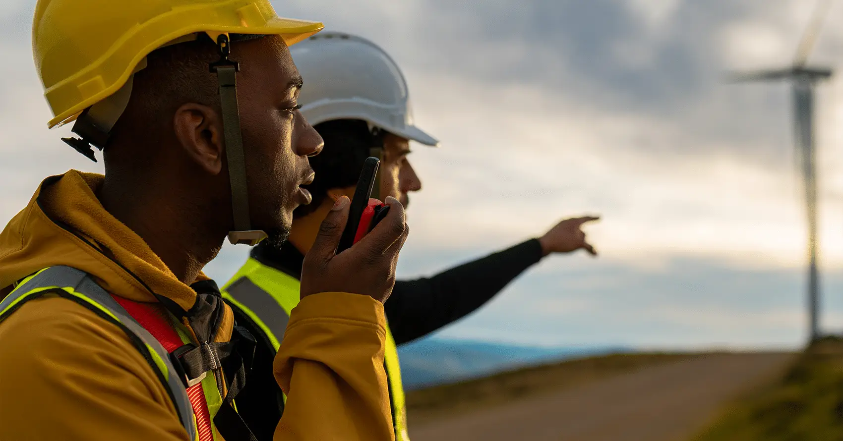 Two construction workers in hard hats and safety vests stand outdoors; one speaks into a walkie-talkie while the other points toward a distant wind turbine.