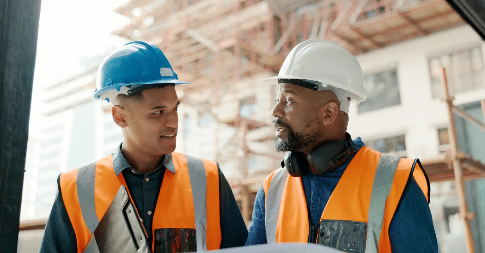 Two construction workers wearing safety vests and hard hats have a discussion at a building site, with scaffolding in the background.