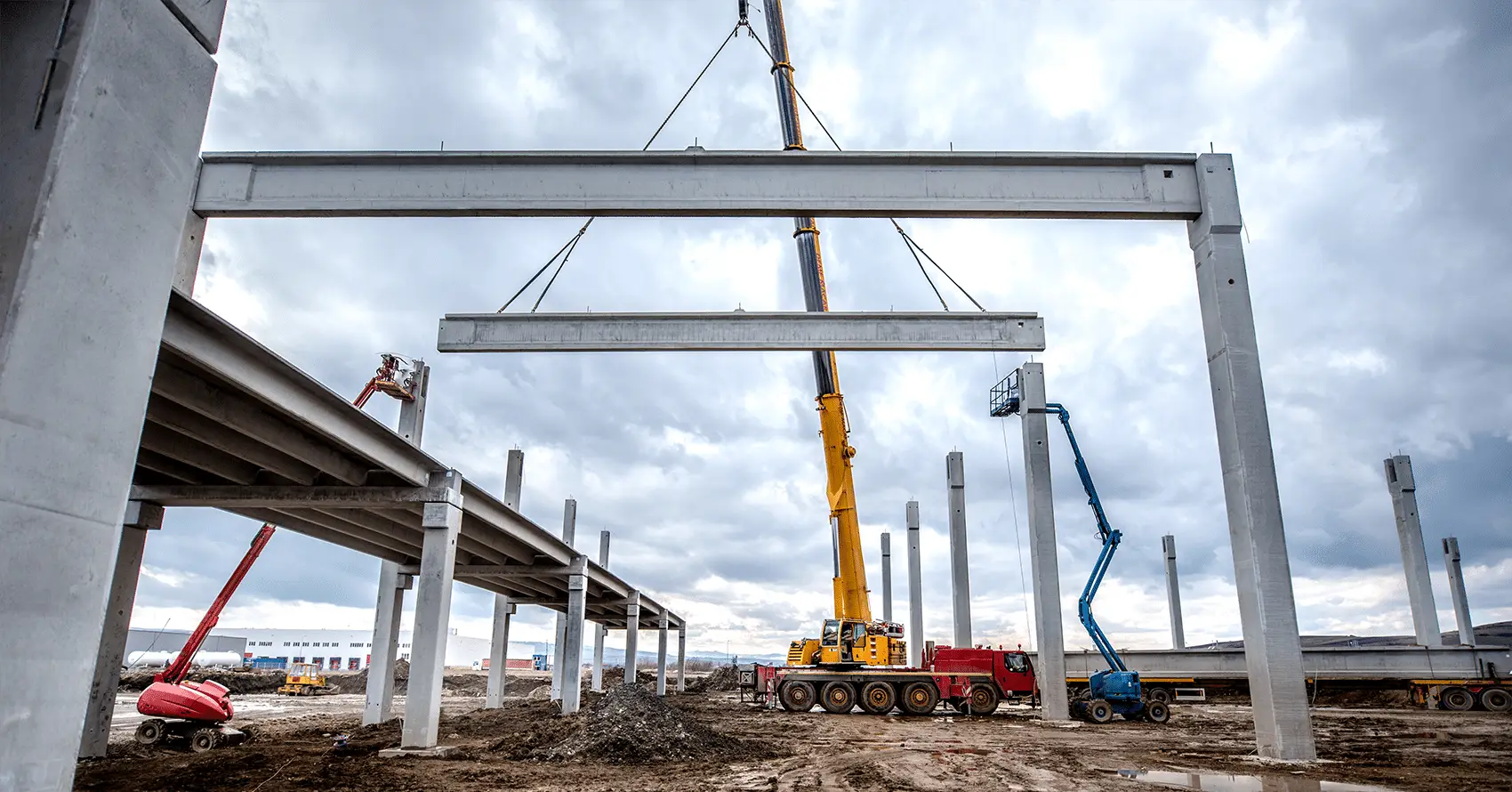 A crane lifts a large concrete beam into place at a construction site, with workers on boom lifts and unfinished building structures visible.