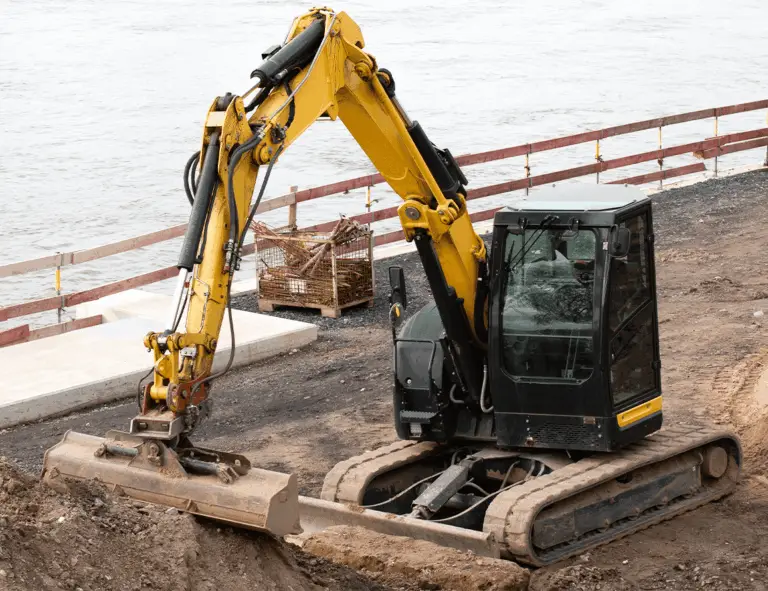 A yellow and black excavator with a bucket attachment is parked on a construction site near water and a wooden safety railing.