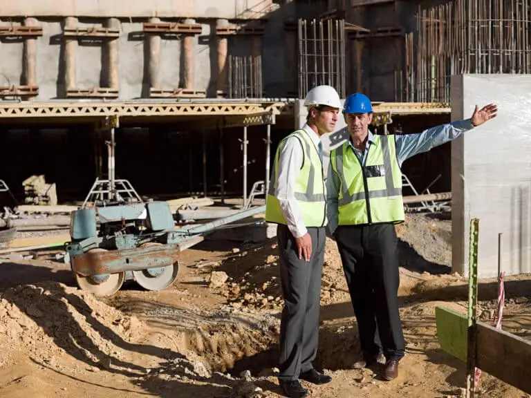 Two men in safety vests and hard hats stand at a construction site, with one man gesturing toward the building structure in the background.