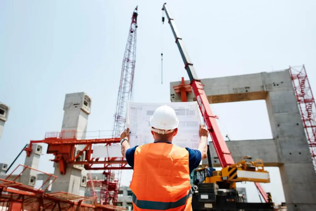 A construction worker in a hard hat and orange vest holds blueprints while facing cranes and a concrete building under construction.