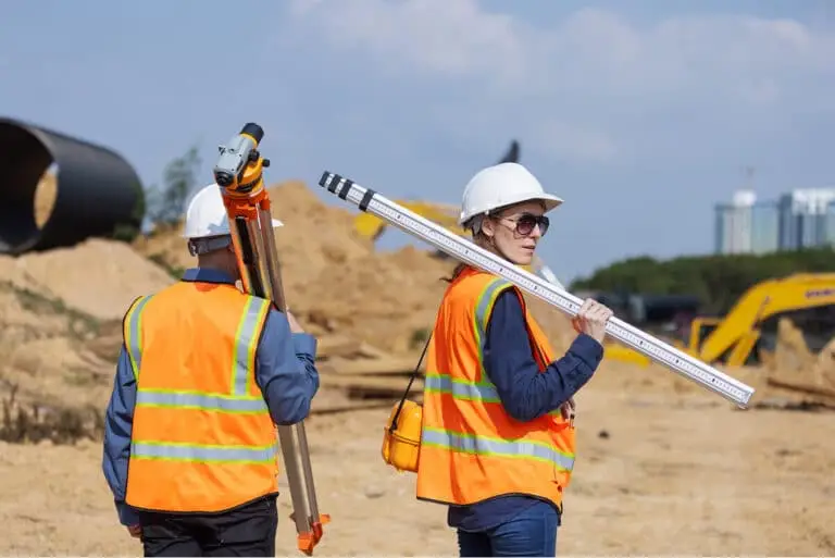 Two construction workers in safety vests and hard hats carry surveying equipment at a construction site with dirt piles and machinery in the background.